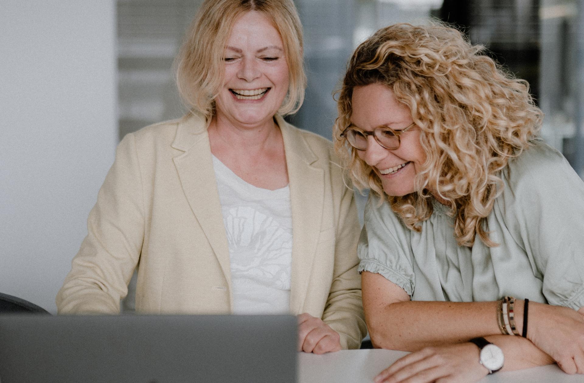 Zwei Frauen beugen sich über einen Laptop und lachen gemeinsam. 
